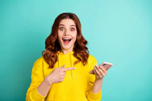 Photo of Photo of beautiful cheerful ecstatic attractive curly wavy trendy stylish youngster pointing at telephone held with hands wearing yellow sweater hoodie isolated over vivid teal color background