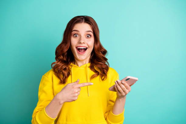 photo of beautiful cheerful ecstatic attractive curly wavy trendy stylish youngster pointing at telephone held with hands wearing yellow sweater hoodie isolated over vivid teal color background - surprise imagens e fotografias de stock