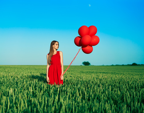 Young beautiful woman in red dress posing in green field with red balloons. Freedom, fun, vacation concept