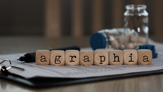 Word AGRAPHIA composed of wooden dices. Pills, documents and a pen in the background. Closeup