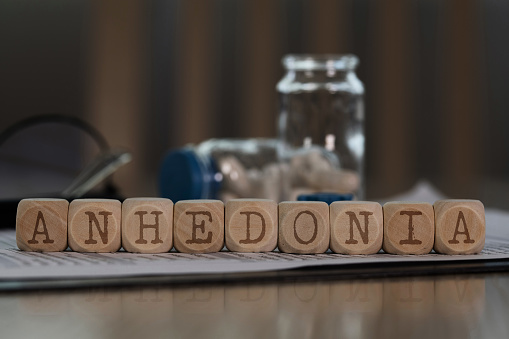 Word ANHEDONIA composed of wooden dices. Pills, documents and a pen in the background. Closeup