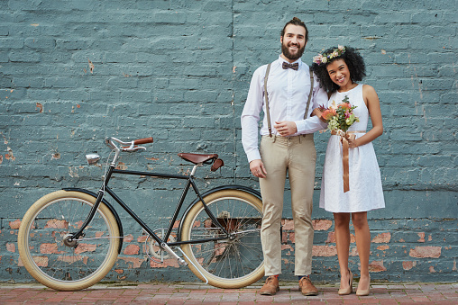 Shot of a newly married young couple standing together against a grey wall with a bicycle next to them