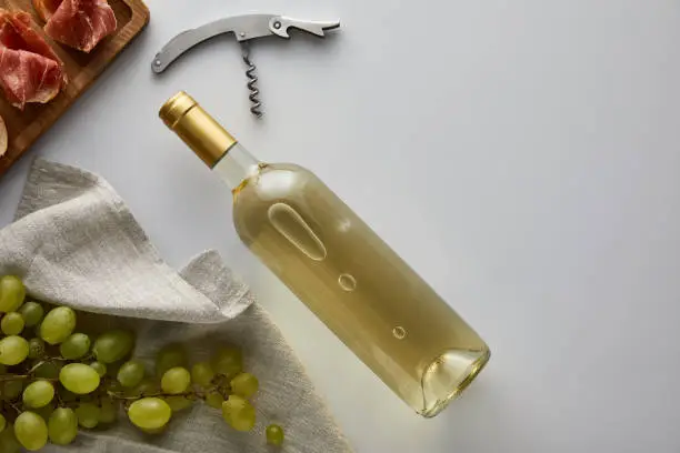 top view of bottle with white wine near grape, corkscrew and sliced prosciutto on baguette on white background