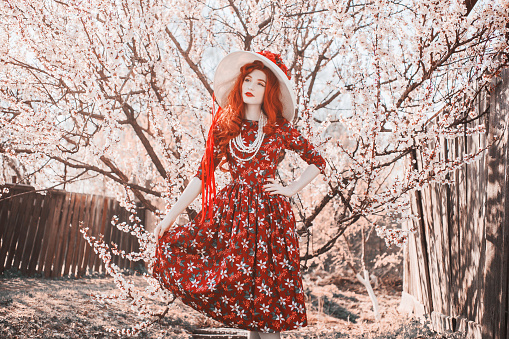 Young beautiful redhead girl with on spring flower garden. Woman with long red hair in retro dress on nature. Blooming spring cherry tree. Stylish girl. Blooming sakura in park. Vanilla flower dress