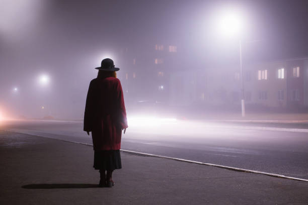 silhouette de femme sur le fond de la ville de nuit dans le brouillard. brume épaisse dans la ville effrayante foncée de soirée. silhouette noire foncée dans le chapeau sur le fond du brouillard. femme seule dans la brume. film de mystère noir. homme  - bad habit fear emotional stress women photos et images de collection