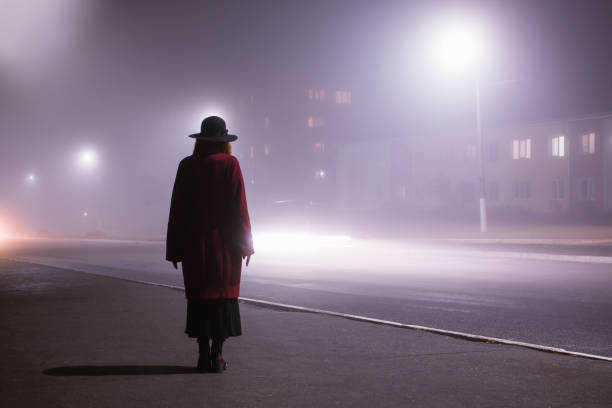 силуэт женщины на фоне ночного города в тумане. густой туман в темном страшном вечернем городе. темный нуар силуэт в шляпе на фоне тумана. од - silhouette women shadow window стоковые фото и изображения