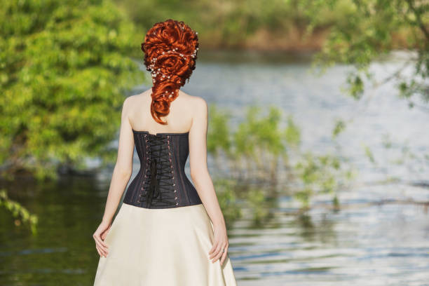 frau zurück. junge viktorianische prinzessin mit frisur auf natur-hintergrund. modell im korsett. rokokokönigin im weißen historischen kleid vor der kulisse von blauem wasser. viktorianische historische nachahmung der gräfin - korsett stock-fotos und bilder