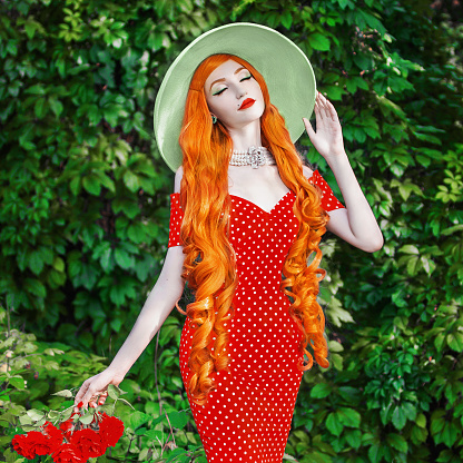 Retro woman in red polka dot dress on green summer background. Vintage redhead girl on background of forest with green leaves. Slim model in hat. Woman holdi branch with roses. Long red hair. Red lips