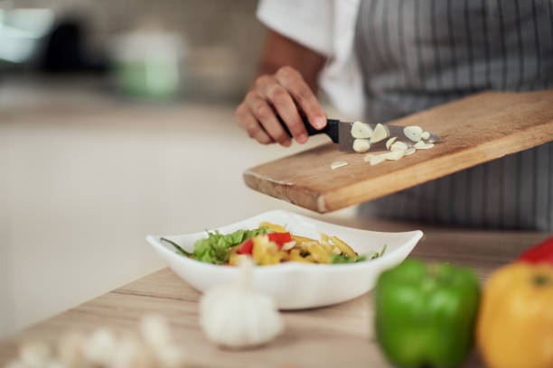 Close up mixed race woman in apron holding cutting board and putting chopped garlic in bowl with vegetables.