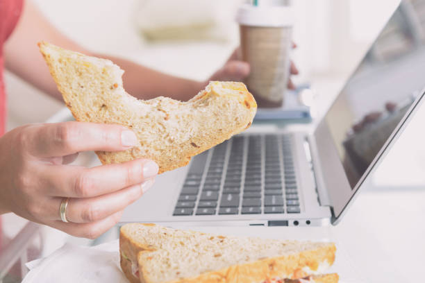 frau, die ein frühstückssandwich isst, während sie mit einem laptop arbeitet - eating sandwich emotional stress food stock-fotos und bilder
