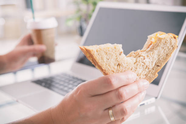 frau, die ein frühstückssandwich isst, während sie mit einem laptop arbeitet - eating sandwich emotional stress food stock-fotos und bilder