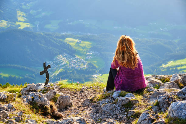 woman near a cross on top of donnerkogel mountain peak, austria, looking towards the green valley below, lit by sunset golden light, at the end of the via ferrata intersport route. - summit cross imagens e fotografias de stock