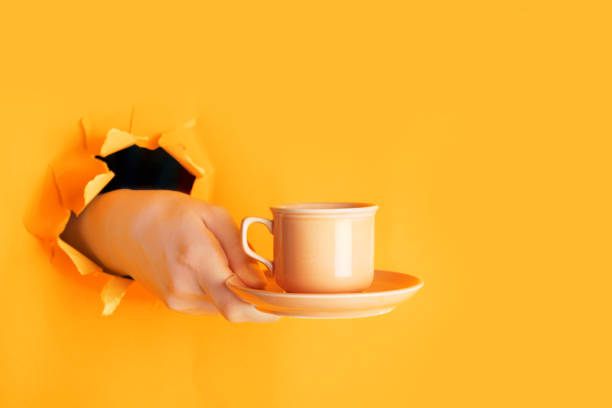 Hand holding a cup of coffee through a hole in torn saffron or light orange paper wall. Coffee break concept, minimalism Hand holding a cup of coffee through a hole in torn saffron or light orange paper wall. Coffee break concept, copy space, minimalism pop art photos stock pictures, royalty-free photos & images