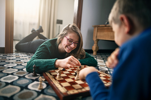 Brother and teenage sister are playing chess on floor. Laughing girl is making a decisive winning move.\nNikon D850