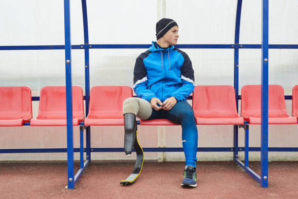 jeune homme paralympique sérieux dans la lame de course utilisant la veste bleue de sports et le chapeau s'asseyant sur des lignes de côté et regardant loin à l'extérieur - blade white blue transportation photos et images de collection