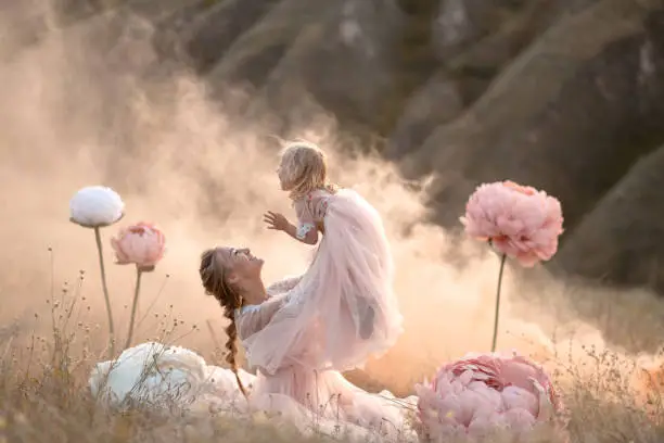 Mom and daughter in pink fairy-tale dresses play in a field surrounded by Big pink decorative flowers.
