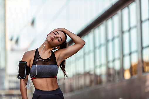 Outdoor shot of beautiful dark hair Caucasian woman athlete wearing black, gray sports bra warming up neck before workout, standing against city with copy space for your promotional content.