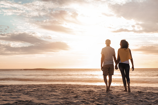 Rearview of a mature couple walking hand in hand together along a sandy beach at dusk