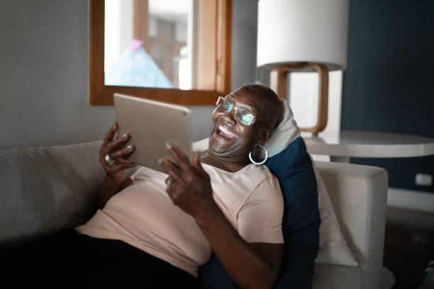 donna anziana sdraiata sul divano usando il tablet - ipad senior adult facebook sofa foto e immagini stock