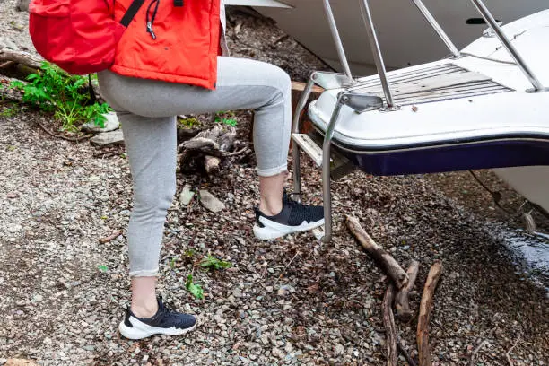 View of the legs of a girl in sneakers who climb aboard a motor boat up the stairs to travel along a river or lake with a guided tour during a trip. Tourism, recreation, sports.
