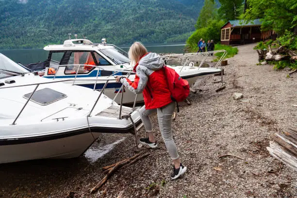 A young blonde woman climbs aboard a motor boat on a ladder to travel along a river or lake with a guided tour during a trip. Tourism, recreation, sports.