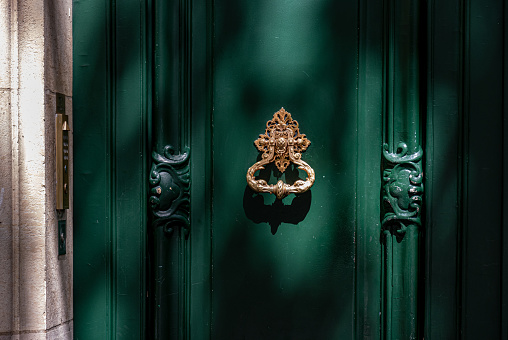 Green painted wooden framed door with gorgeous antique ornate knocker and carving sculptural details. Architectural features of Paris door in bright sunlight with shadows at old building in France.