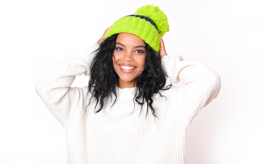 Close-up photo of alluring African American woman in snow-white sweater and green hat with pompom, who is posing with her hands on her head and smiling irresistibly.