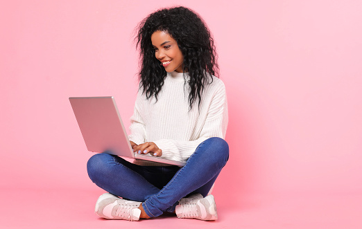 Hit the ground running. Magnificent African American lady in a snow-white sweater is sitting cross-legged with her laptop, smiling happily while looking at the screen.