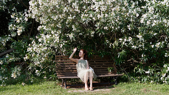 Portrait of beautiful Chinese young girl in green dress sitting in park wooden bench with hand protecting sunlight under trees full of white flowers.