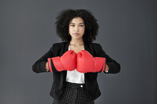Studio portrait of a young corporate businesswoman posing wearing boxing gloves against a grey background
