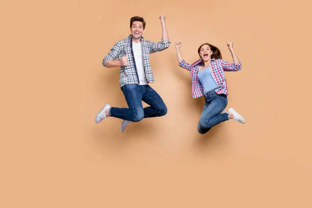 Photo of Full length photo of two people crazy lady guy jumping high celebrating best win raising fists excited wear casual plaid jeans clothes isolated beige background