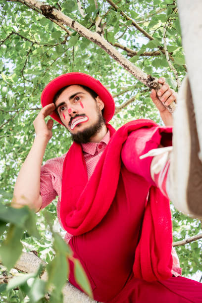 un mimo triste está sentado en un árbol con ropa roja. payaso pensando en un abedul. - face paint human face mask carnival fotografías e imágenes de stock