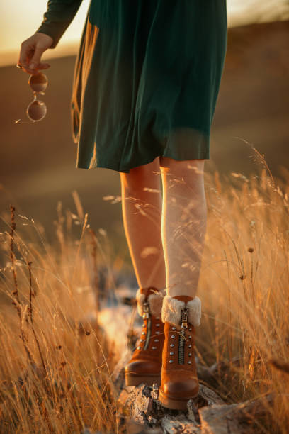 Attractive young woman walking over tree trunk on meadow. Sunset light makes colors and light looks warm, soft and beautiful. She wears green dress and boots.