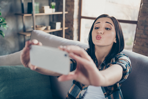 Photo of charming lady holding telephone making selfies speaking, skype sending boyfriend air kisses sit comfort sofa wear casual checkered shirt apartment indoors