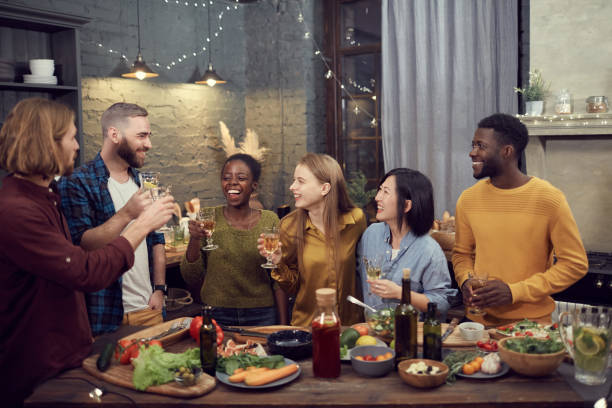 Diverse Group of Young People Enjoying Dinner Party Multi-ethnic group of smiling young people enjoying dinner together standing at table in modern interior and holding wine glasses, copy space people banque stock pictures, royalty-free photos & images
