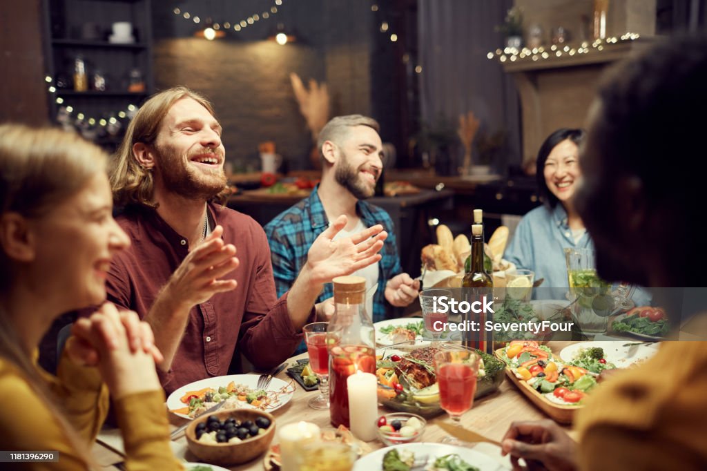 People Laughing at Dinner Table Group of emotional young people enjoying dinner party with friends and smiling happily sitting at table in dimly lit room, copy space Friendship Stock Photo