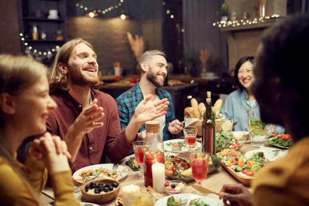 People Laughing at Dinner Table Group of emotional young people enjoying dinner party with friends and smiling happily sitting at table in dimly lit room, copy space people banque stock pictures, royalty-free photos & images