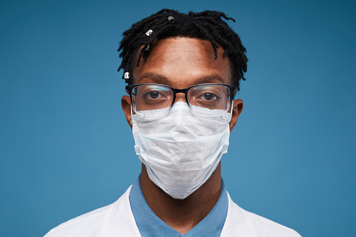 Head and shoulders portrait of young African-American doctor wearing protective mask and looking at camera while posing against blue background