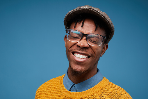 Head and shoulders portrait of excited African-American man smiling happily while posing against blue background wearing bright knit sweater