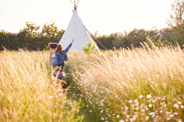 Two Female Friends Walking Pulling Trolley Through Field Towards Teepee On Summer Camping Vacation Two Female Friends Walking Pulling Trolley Through Field Towards Teepee On Summer Camping Vacation glamping photos stock pictures, royalty-free photos & images