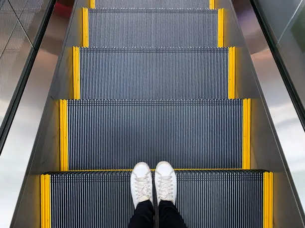 Selfie of feet in white sneakers shoes standing on escalator in shopping mall or modern office