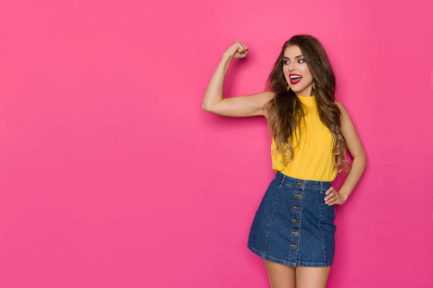 Happy Young Woman In Jeans Mini Skirt And Yellow Top Is Flexing Biceps And Talking stock photo