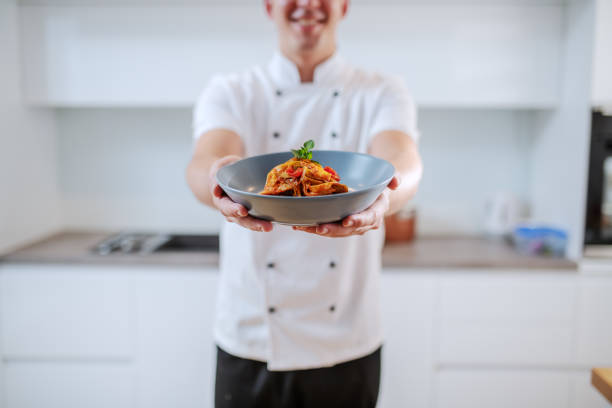 istock Smiling caucasian creative chef in white uniform holding plate with tagliatelle while standing in kitchen. Selective focus on plate. 1181448318