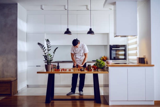 Full length of handsome caucasian creative chef standing in kitchen and cutting salmon for lunch. On kitchen counter are vegetables and spices.