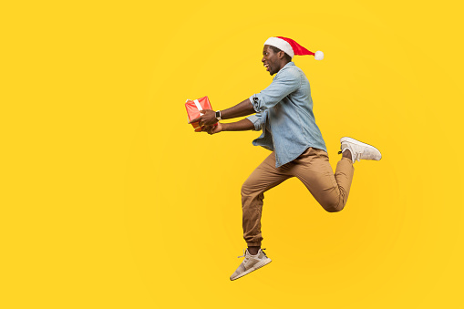 Full length side view of joyous handsome young man in santa hat and casual denim shirt jumping or running with christmas gift box, hurry for sales.