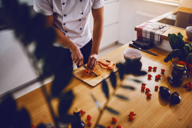 Caucasian creative chef standing in kitchen and cutting salmon for lunch. On kitchen counter are vegetables and spices.