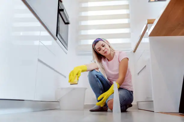 Worthy caucasian blonde housewife with rubber gloves on hands holding sponge and cleaning floor while crouching in kitchen. Next to her is bucket.