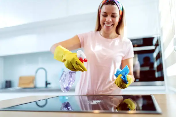 Worthy cheerful caucasian blond housewife with rubber gloves on hands spraying stove and cleaning it. Kitchen interior.