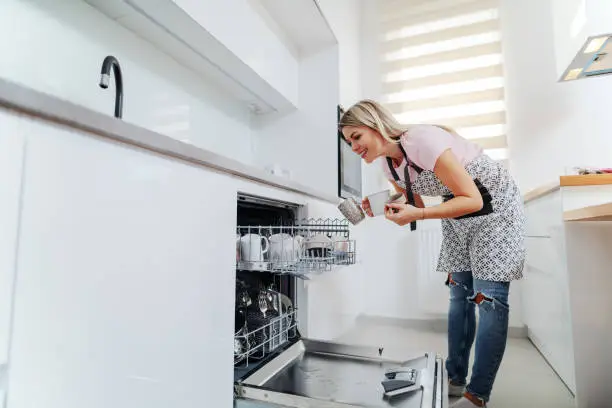 Worthy caucasian housewife in apron putting mugs in dishwasher while standing in kitchen.