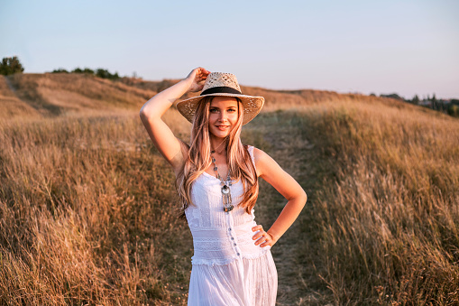 Beautiful woman wearing in white dress and straw hat enjoying nature outdoors at sunset.